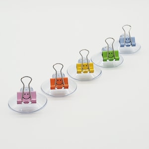 Puede incluir: Cinco clips de ventosa con caritas sonrientes. Los clips son de diferentes colores: rosa, naranja, amarillo, verde y azul.