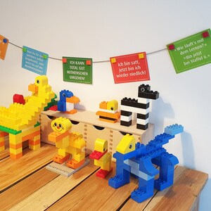 May include: A collection of colorful Lego animal figures, including a yellow duck, a blue penguin, a yellow giraffe, a yellow and orange lion, a yellow duck, a black and white zebra, and a blue dinosaur. The figures are arranged on a wooden table with a string of colorful cards hanging above them. The cards have German text on them.