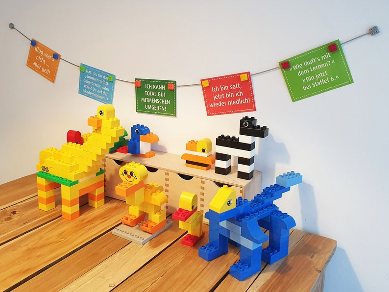 May include: A collection of colorful Lego animal figures, including a yellow duck, a blue penguin, a yellow giraffe, a yellow and orange lion, a yellow duck, a black and white zebra, and a blue dinosaur. The figures are arranged on a wooden table with a string of colorful cards hanging above them. The cards have German text on them.