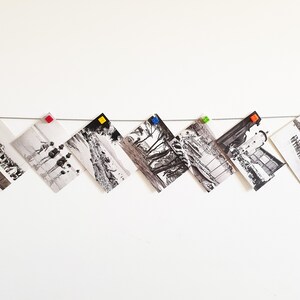 May include: A white wall with a string of black and white photos hanging from it. The photos are held in place by colorful plastic clips. The photos are all different sizes and shapes.