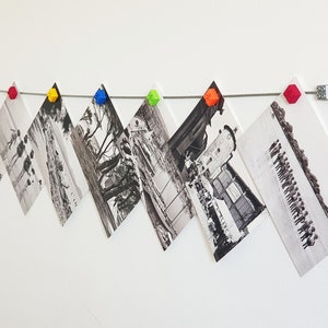 May include: A string of black and white photographs are hung on a white wall with colorful plastic clips. The photos are arranged in a staggered pattern.