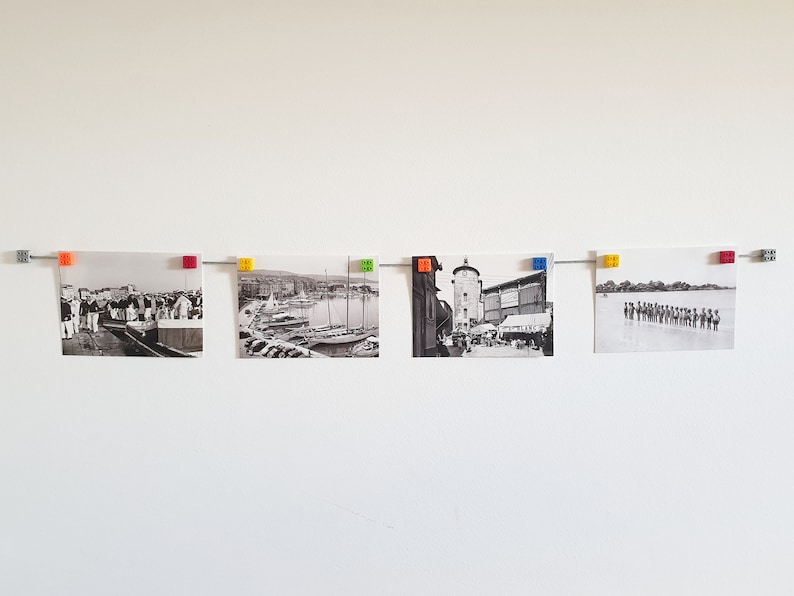 May include: Five black and white photographs are displayed on a white wall using colorful plastic clips. The photos show various scenes, including a harbor with boats, a city street, and a group of people.