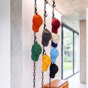 Peut inclure: Trois chaînes métalliques noires verticales suspendues au plafond, chacune dotée de plusieurs crochets intégrés. Dix casquettes de baseball de différentes couleurs unies, dont orange, vert, gris foncé, jaune, rouge, crème, bleu et noir, y sont suspendues.