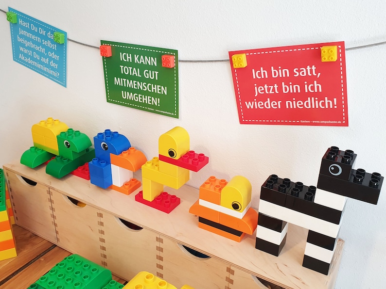 May include: A collection of colorful toy animals made from large plastic building blocks. The animals include a green turtle, a blue bird, a yellow duck, an orange and yellow fish, and a black and white zebra. The animals are arranged on a wooden shelf. The shelf is in front of a white wall with three signs hanging on a string. The signs have text in German. The text on the signs reads: "Hast Du Dir da Jammern selbst beigebracht, oder warst Du auf der Akademimimimi?", "ICH KANN TOTAL GUT MITMENSCHEN UMGEHEN!" and "Ich bin satt, jetzt bin ich wieder niedlich!"