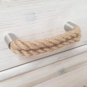 Pode incluir: Um puxador de armário com uma pega de corda natural e extremidades de metal escovado. A corda é de cor castanho claro e as extremidades metálicas são prateadas. O puxador é montado numa superfície de madeira pintada de branco.