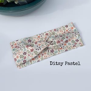 Pu&ograve; includere: Una fascia floreale con un design a nodo attorcigliato. Il tessuto presenta un motivo di piccoli fiori in colori pastello, tra cui rosa, blu e pesca, su uno sfondo crema. Il testo "Ditsy Pastel" &egrave; stampato sull'immagine.