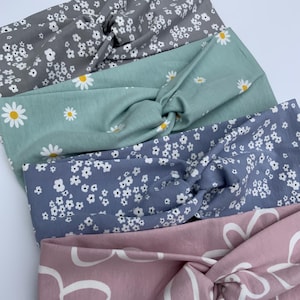 Pu&ograve; includere: Quattro fasce per capelli in tessuto, con un design a nodo attorcigliato, nei toni del grigio, verde acqua, blu e rosa. Le fasce sono decorate con motivi floreali, tra cui margherite bianche e piccoli fiori bianchi. La fascia rosa ha un grande motivo floreale bianco.
