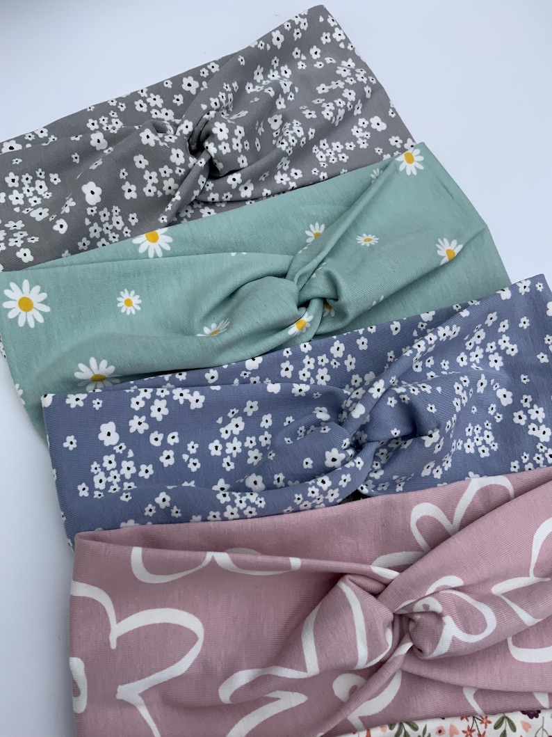 Pu&ograve; includere: Quattro fasce per capelli in tessuto, con un design a nodo attorcigliato, nei toni del grigio, verde acqua, blu e rosa. Le fasce sono decorate con motivi floreali, tra cui margherite bianche e piccoli fiori bianchi. La fascia rosa ha un grande motivo floreale bianco.