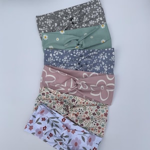 Pu&ograve; includere: Un assortimento di sette fasce per capelli floreali in vari colori, tra cui grigio, verde salvia, lavanda e rosa. Ogni fascia presenta un design annodato ed &egrave; adornata con diversi motivi floreali, come margherite e fiori stilizzati.