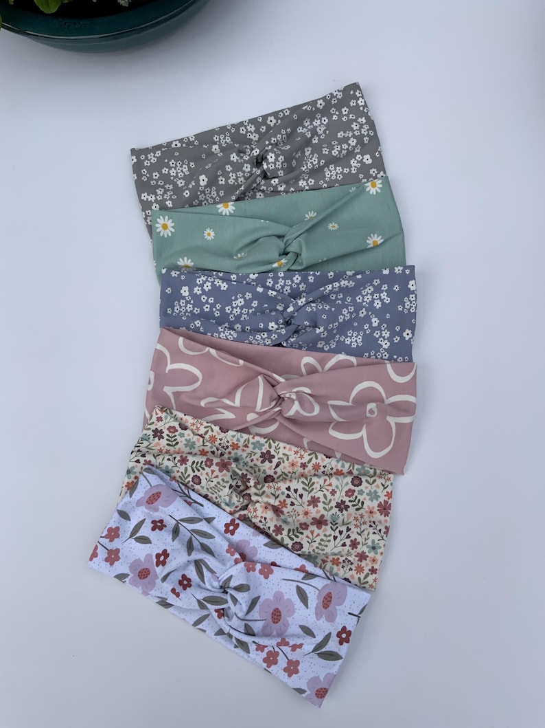 Pu&ograve; includere: Un assortimento di sette fasce per capelli floreali in vari colori, tra cui grigio, verde salvia, lavanda e rosa. Ogni fascia presenta un design annodato ed &egrave; adornata con diversi motivi floreali, come margherite e fiori stilizzati.
