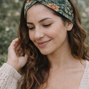 Pu&ograve; includere: Una fascia floreale verde con un design annodato. La fascia presenta un motivo di fiori e foglie arancioni e gialle. La donna nell'immagine indossa un maglione beige e ha lunghi capelli castani ondulati.