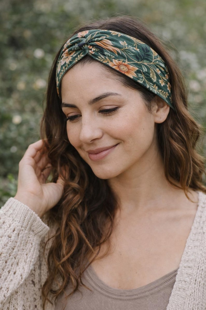 Pu&ograve; includere: Una fascia floreale verde con un design annodato. La fascia presenta un motivo di fiori e foglie arancioni e gialle. La donna nell'immagine indossa un maglione beige e ha lunghi capelli castani ondulati.