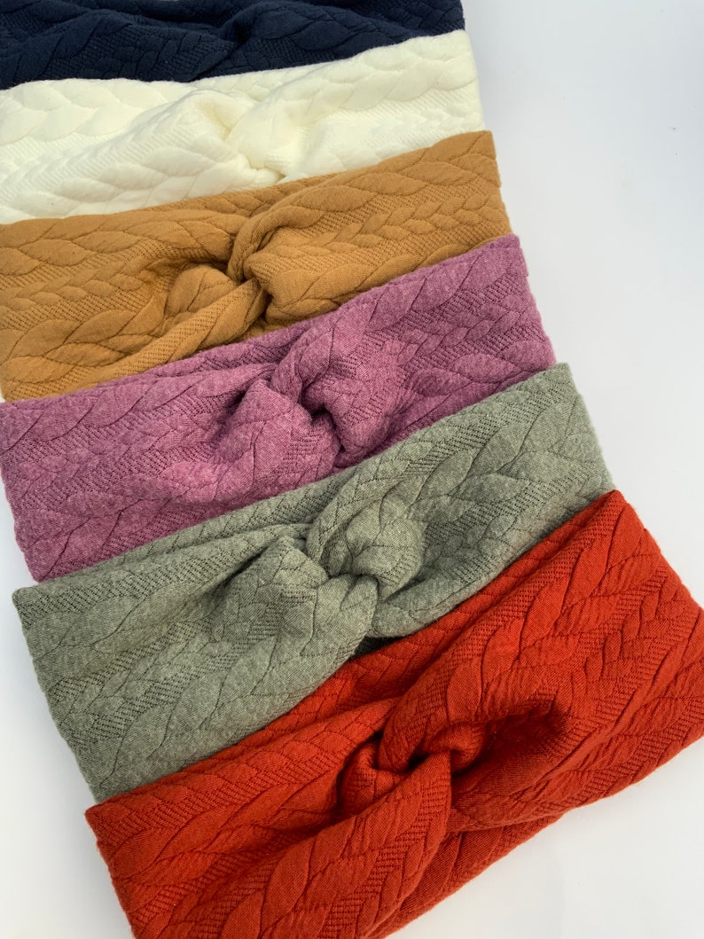 Puede incluir: Seis diademas de punto de diferentes colores con un patr&oacute;n de punto trenzado. Las diademas est&aacute;n apiladas una encima de la otra, con el color m&aacute;s oscuro en la parte superior y el color m&aacute;s claro en la parte inferior. Los colores son azul marino, blanco, marr&oacute;n, morado, verde y naranja.