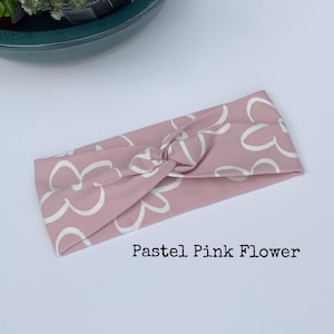 Pu&ograve; includere: Una fascia per capelli rosa pastello con un motivo floreale bianco. La fascia &egrave; annodata al centro. Il testo "Pastel Pink Flower" &egrave; stampato sotto.