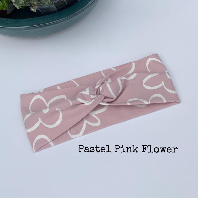 Pu&ograve; includere: Una fascia per capelli rosa pastello con un motivo floreale bianco. La fascia &egrave; annodata al centro. Il testo "Pastel Pink Flower" &egrave; stampato sotto.