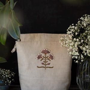 Op de afbeelding: Een beige tas met trekkoord voorzien van een centraal borduurwerk van een gestileerde bloem met mauve bloemblaadjes, witte accenten en groene stengels. De tas is gemaakt van een textuurrijke stof en heeft een sluiting met geknoopte uiteinden.