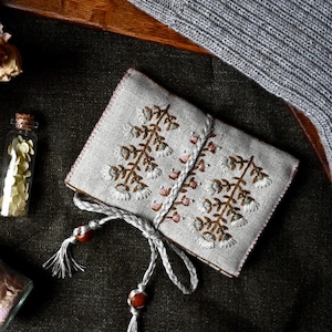 May include: A light grey embroidered pouch with a floral design in brown and white. The pouch is tied with a grey cord and wooden beads. The background includes a grey knitted item and small glass bottles.