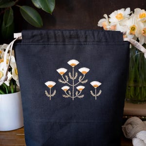 May include: A black fabric tote bag with a drawstring closure. The bag features an embroidered floral design in white and gold. The bag is shown with white flowers in a vase and a potted plant in the background.