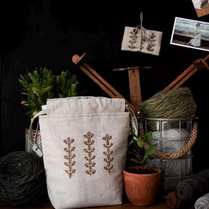 May include: A beige linen yarn bag with three embroidered floral stems. The bag sits on a wooden surface with other craft supplies, including yarn, a small potted plant, and a wooden yarn swift. A small embroidered item hangs in the background.