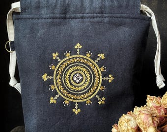 Puede incluir: Una bolsa azul marino con cordón y bordado floral dorado. La bolsa presenta un diseño circular con una flor central, cordones blancos y un pequeño anillo de metal.