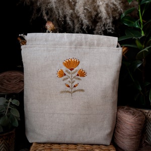 Peut inclure: Un sac fourre-tout en lin beige avec une broderie florale orange et marron. Le sac rectangulaire présente un design simple et est posé sur un panier en osier. Idéal pour le rangement.