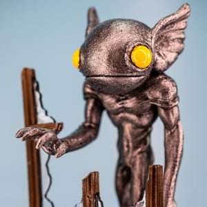 Op de afbeelding: Een metalen beeldje van een wezen met grote gele ogen en vleugelachtige oren, naast een gebroken houten raamkozijn. Het figuur heeft een humanoïde vorm met gedetailleerde texturen en een bronzen afwerking. Het raamkozijn is bruin met een zilverkleurig gebroken ruit.