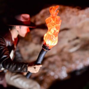 May include: A person wearing a brown leather jacket and a hat holds a torch with a bright orange flame. The torch is made of wood and has a metal tip.
