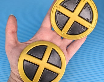 May include: Two gold-colored round coasters with a black X-shaped design in the center. The coasters have a raised edge and a smooth surface.