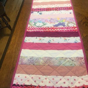 May include: A pink and white quilted table runner with a variety of patterned fabric strips. The runner features hearts, polka dots, and floral designs.