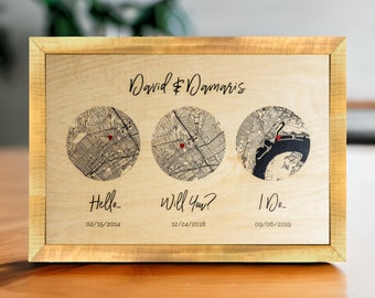 Può includere: Una stampa incorniciata in legno con tre mappe della stessa città. Ogni mappa ha uno spillo rosso a forma di cuore che segna una posizione diversa. Il testo "David & Damaris" è in cima alla stampa. Sotto le mappe ci sono le date "02/15/2014", "12/24/2018" e "09/06/2019" con i testi "Hello...", "Will You?" e "I Do..." rispettivamente.