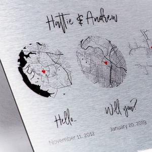 Peut inclure: Une plaque en métal argentée avec le texte "Hattie & Andrew" en haut. Deux cartes en noir et blanc avec des cœurs rouges marquant des emplacements sont affichées. Le texte "Hello... November 11, 2012" est sous la première carte et le texte "Will you? January 20, 2019" est sous la deuxième carte.