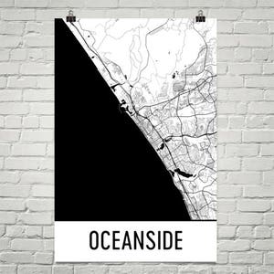 Puede incluir: Un mapa en blanco y negro de Oceanside, California, con el nombre de la ciudad en texto blanco en negrita en la parte inferior. El mapa muestra la costa y las calles de la ciudad. El póster está colgado en una pared de ladrillos blancos.