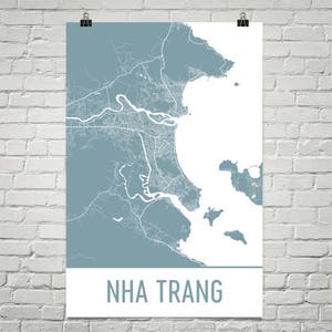 Op de afbeelding: Een poster met een gedetailleerde kaart van Nha Trang, Vietnam, in blauw- en wittinten. De kaart toont de stadsindeling, de kustlijn en de waterwegen. De naam van de stad staat onderaan in een vetgedrukte lettertype.