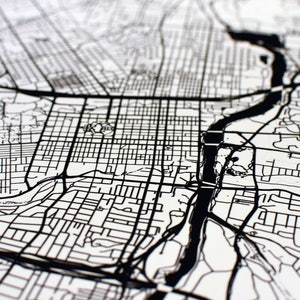 May include: Black and white detailed map print. The map features a river, roads, and city blocks. The map is printed on a white background. The image is a close-up view of the map.