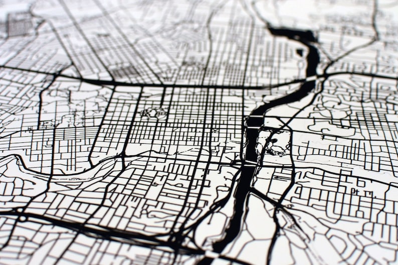May include: Black and white detailed map print. The map features a river, roads, and city blocks. The map is printed on a white background. The image is a close-up view of the map.
