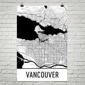 May include: A black and white map of Vancouver, Canada, displayed on a white poster. The map details the city's streets, waterways, and surrounding areas. The word "VANCOUVER" is printed in bold, black letters at the bottom of the poster.