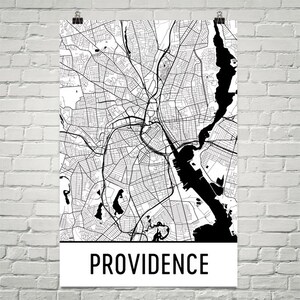May include: Black and white map of Providence, Rhode Island, featuring intricate street and waterway details. The city map is printed on a white background with the city name in bold, black letters at the bottom. The map is hanging on a white brick wall.