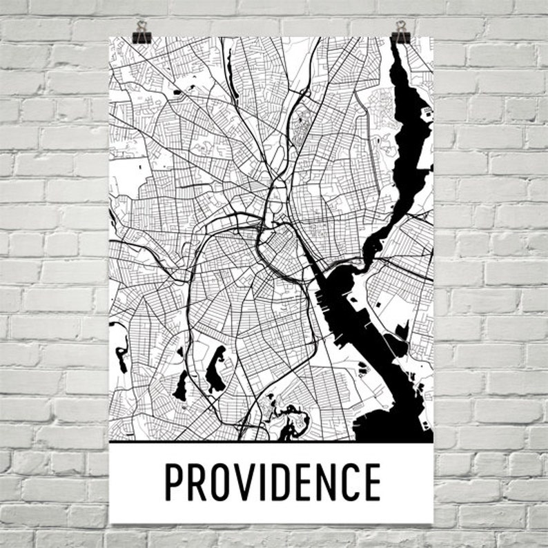 May include: Black and white map of Providence, Rhode Island, featuring intricate street and waterway details. The city map is printed on a white background with the city name in bold, black letters at the bottom. The map is hanging on a white brick wall.