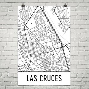 May include: A black and white map of Las Cruces, New Mexico, USA, with a detailed street layout. The city name is printed in bold, black letters at the bottom of the poster. The map is printed on white paper and is held by two black binder clips.