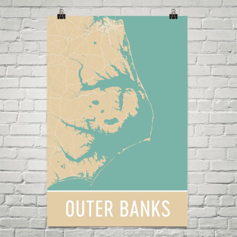 May include: A poster featuring a map of the Outer Banks in North Carolina. The map is in shades of beige and teal, with the text "Outer Banks" in a beige banner at the bottom. The poster is held by a black clip against a white brick wall.