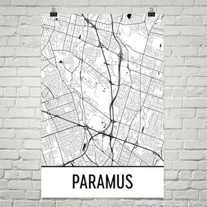 May include: A monochrome map of Paramus, New Jersey, showing the road and street layout. The town's name is printed in bold, black letters at the bottom of the poster. The map is displayed on a white background and is held by two silver clips.