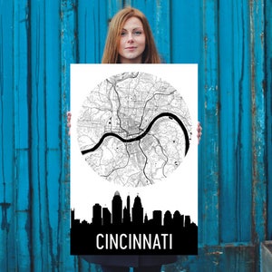 May include: A black and white print featuring a map of Cincinnati, Ohio, in a circular design. Below the map is a black silhouette of the city's skyline, with the city name in white text. The print is held by a person.