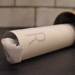 May include: A white poster tube with a gray "R" printed on the side. The tube is partially unrolled and has a brown cardboard end cap.