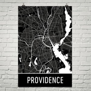 May include: A black and white map of Providence, Rhode Island, with a detailed street layout. The city's name is displayed in white block letters on a black banner at the bottom of the poster. The map is hanging on a white brick wall.