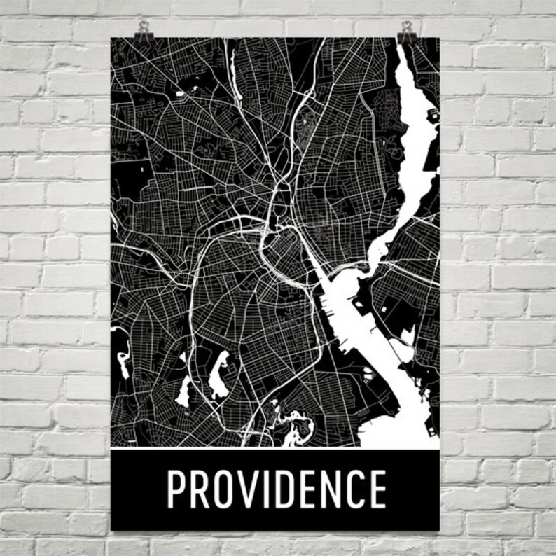 May include: A black and white map of Providence, Rhode Island, with a detailed street layout. The city's name is displayed in white block letters on a black banner at the bottom of the poster. The map is hanging on a white brick wall.