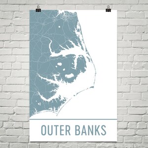 May include: A poster featuring a map of the Outer Banks in a muted blue-gray color. The map shows the coastline and roads in white against the blue background. The words "Outer Banks" are printed in a matching blue-gray font at the bottom.