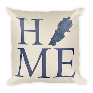 Op de afbeelding: Cr&egrave;mekleurig vierkant kussen met het woord "HOME" in marineblauwe blokletters. Een marineblauwe kaart van Libanon staat boven de letter "ME". Het kussen heeft een zachte, uitnodigende uitstraling.