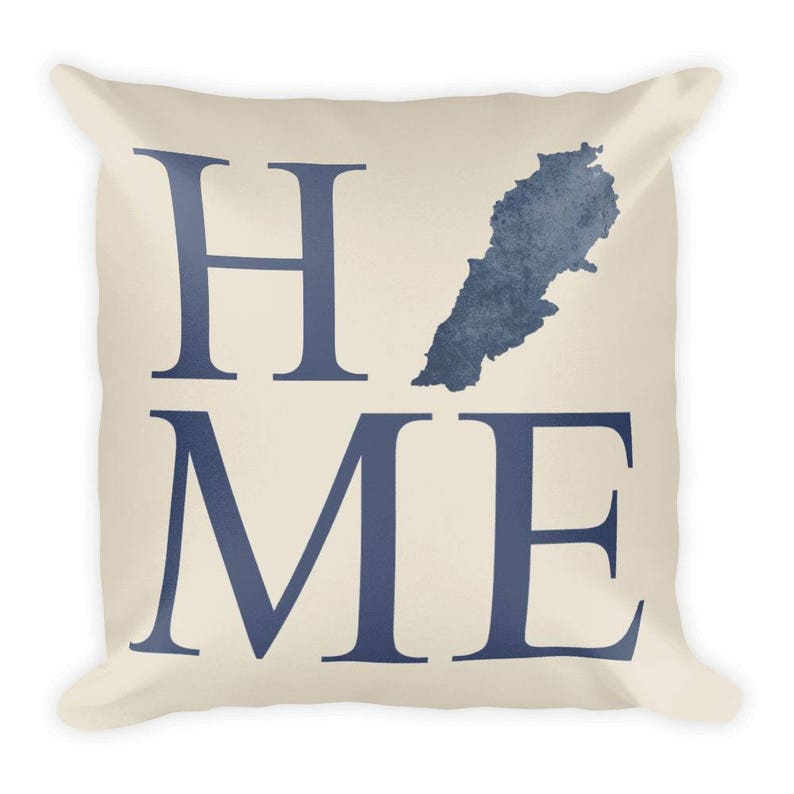 Op de afbeelding: Cr&egrave;mekleurig vierkant kussen met het woord "HOME" in marineblauwe blokletters. Een marineblauwe kaart van Libanon staat boven de letter "ME". Het kussen heeft een zachte, uitnodigende uitstraling.