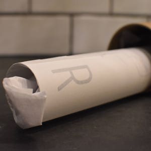 May include: A white poster tube with a gray "R" printed on the side. The tube is partially unrolled and has a brown cardboard end cap.