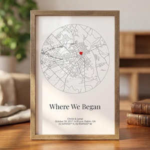 Op de afbeelding: Een ingelijste print met een cirkelvormige kaart en een rode markering. De tekst "Where We Began" staat onder de kaart, samen met namen, een datum, tijd en coördinaten. De lijst is lichtbruin.